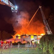 Flames and smoke engulf a large house in Maryland as a NIHFD ladder hovers over, with a fireman up top spraying water from a hose