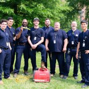 Eight guys stand smiling on the lawn, one with his foot resting on a generator