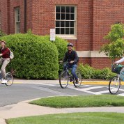Three cyclists on bikes, wearing helmets, round the corner of parking lot toward the pit stop.