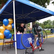 A cyclist hunches over upside-down bike looking in front of NIH Police tent, yellow and blue balloons