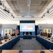 Tabak and Sadikin sit at desk in center of room with slide show presentation behind them. Rows of other attendees face center of room.