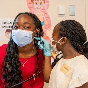 Girl in white shirt, right, uses an otoscope to examine the ear of her sister, left, in red shirt.
