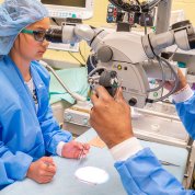 Child and adult in surgical caps and gowns peer into microscope.