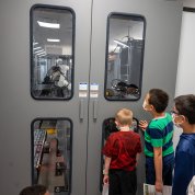 Three masked children peer through windows at a robotic arm.