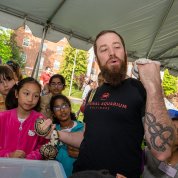 Man with snake tattoo holds up two live snakes for crowd of children.