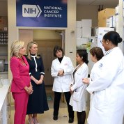 Several women--four in labcoats--chat in a lab environment with NIH NCI logo on wall behind them.