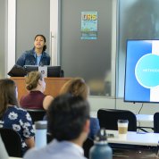 Woman at lectern beside screen, with students at desks in foreground