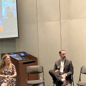 Two women and a man sitting in chairs with the title screen projected behind them