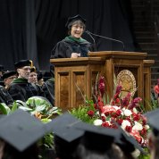 Bertagnolli in black cap and gown stands at podium as graduates wearing caps look on.
