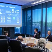 large conference table with several men and women seated around it. One man stands at the lectern, gesturing toward the slide projected beside him.
