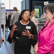 Woman gestures with hands as she speaks with another woman, with posters all around them.