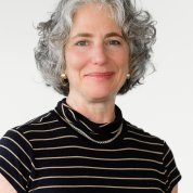 Ascher smiles against a plain white background. She has short gray hair and is wearing a black striped shirt.