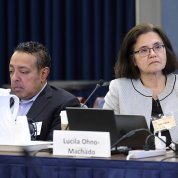 At conference table, a man looks down at his computer, a woman watches the projection screen.