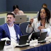At a conference table, a man watches the projection screen as a woman seated beside him speaks, gesturing with her hand.