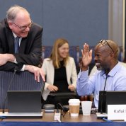 One man standing and another seated at the conference table smile at each other