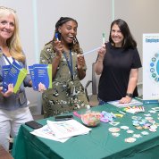 Three smiling women hold pens and brochures next to Employee Assistance Program sign.