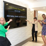 NIH director and two event organizers smile while pointing to Wellness Day sign on large TV screen.