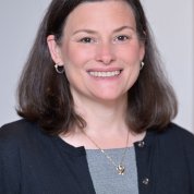 Cernich, wearing a gray shirt and black cardigan, smiles against a gray backdrop.