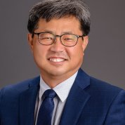 Jeong, wearing a suit and tie, smiles against a dark gray backdrop.