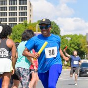 A smiling runner, wearing a blue outfit and black hat, passes a group of onlookers. The runner holds a yellow baton.