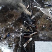 looking down at train cars tilted and off the tracks, large plume of gray and black smoke at top of frame, other wreckage nearby