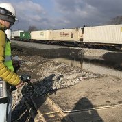 man in hardhat and insulated yellow safety jacket holds instruments close to train tracks 