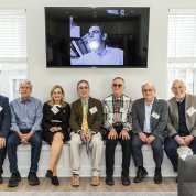 Several men and a woman sitting side by side on the long-bench window seat, with windows and a screen projecting Leder's photo behind them