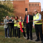 A line of people stand behind a multicolored ribbon. Three people wear suits and three wear fluorescent vests.