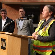 A woman in a fluorescent vest speaks into a microphone at a podium bearing an NIH logo. Two men look on in the background.