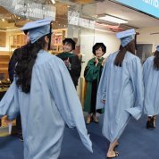 In entryway, graduates in caps and gowns walk past Bertagnolli and Fried who are clapping.