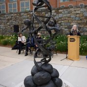 Statue of DNA double helix with four infants ascending it. Several seated individuals and one at a podium look on in the background.