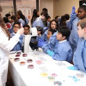 Group of kids, some looking in awe, others looking grossed out, stand around table as a tech holds up a petri dish of bacteria.