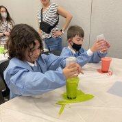 Two kids pour chemicals into a glass, one looking neon green, the other peach, during an experiment.