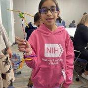 A girl in an NIH sweatshirt holds up pipe cleaners in the shape of a neuron.