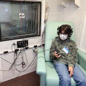 A child sits with headphones with protruding wires holding a signaling device in front of window inside booth.