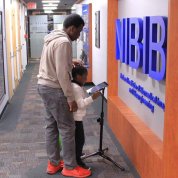 A man stands behind a child and video tablet, with NIBIB on wall, in a corridor of Bldg. 31.
