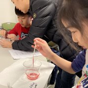 A young child holds a stick into a cup of mushy strawberry at a table.