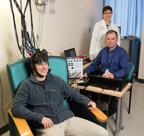 A young man sits smiling with a helmet of wires, as two doctors look on.