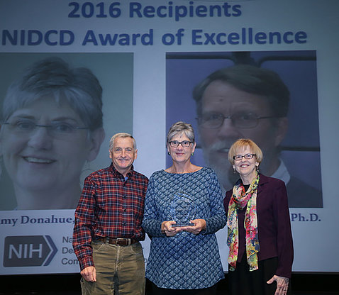 Donahue stands on stage, holding award, with, Battey and Cooper in front of Award of Excellence slide 