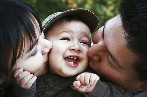 Young child grins with several teeth showing, is kissed on either side by a parent