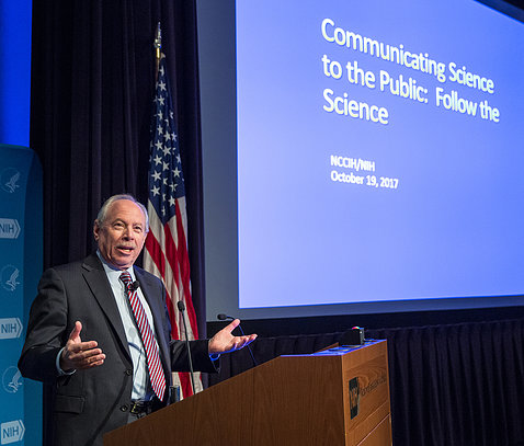 Leshner speaks with hands open at podium, next to slide that reads: Communicating Science to the Public