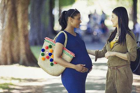 Two women of color, one holding her pregnant belly, chat in park