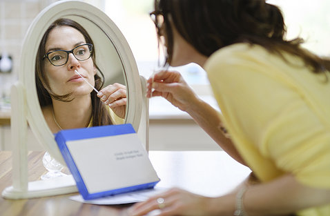 A woman sits in front of a mirror about to insert a long testing swab into her nose.