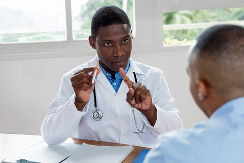Black male doctor holds cigarette in one hand and wags finger with other hand to warn black male patient not to smoke.