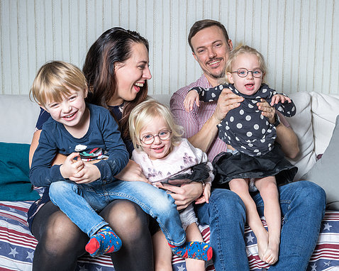 Niclas and Jessica, with their 3 young children, all smiling, sit together on a bed