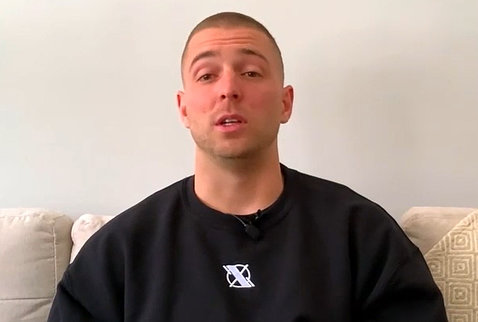 A screenshot of Thompson, wearing a black sweatshirt, seated on his couch at home, speaking on videocast.