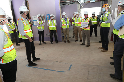About a dozen people in helmets, masks, and safety vests stand in an unfinished room as one speaks.