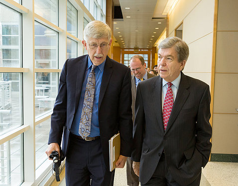 Collins, holding a notebook, walks down a hallway at NIH talking with Sen. Blunt