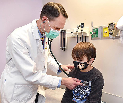 A doctor in a white lab coat holds a stethoscope to a child's chest in his office.