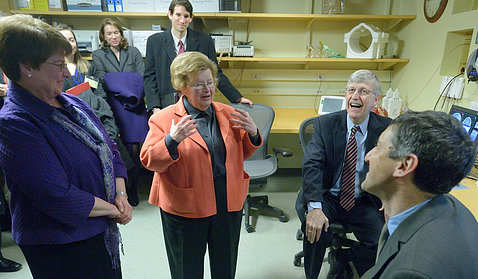 Former Maryland Senator Barbara Mikulski tells a story standing in an NIH lab as Collins seated nearby laughs.